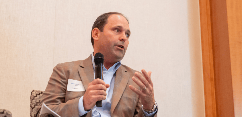 A professional in a brown suit, Brandon Moorefield articulates a strategic vision into a microphone, using confident gestures to engage his audience.