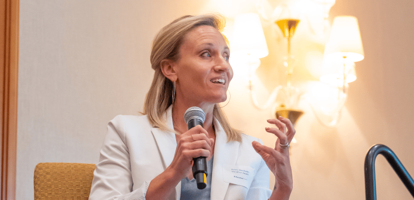 Krista Beckwith speaks into a microphone at a roundtable, gesturing with her right hand while seated.