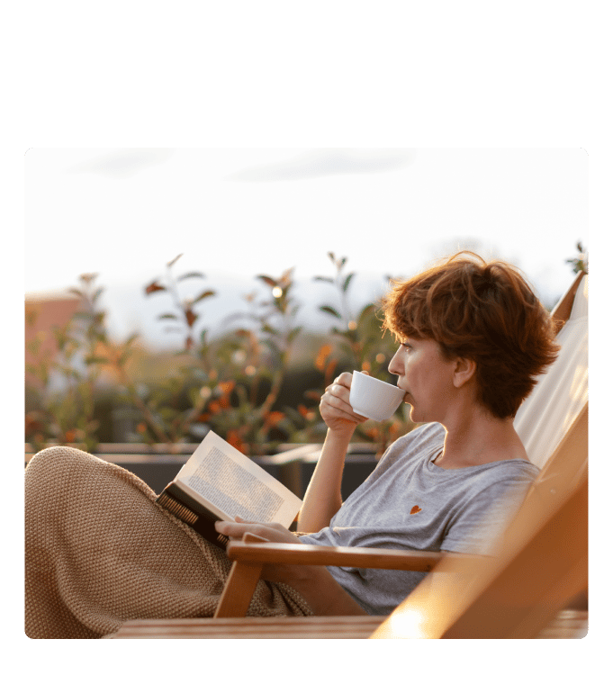 Woman sipping from a cup while reading a book on a terrace.