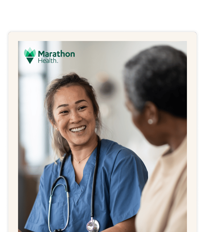 A smiling Marathon Health nurse in blue scrubs talks warmly with an older female patient during a consultation.
