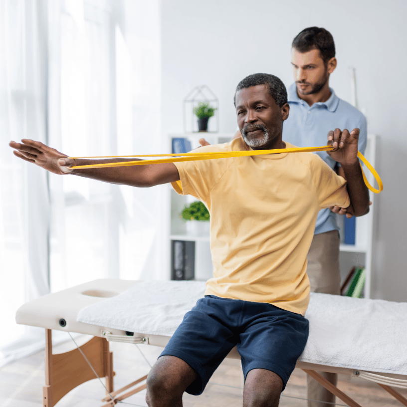 An older man performs a resistance band exercise while a physical therapist assists from behind.