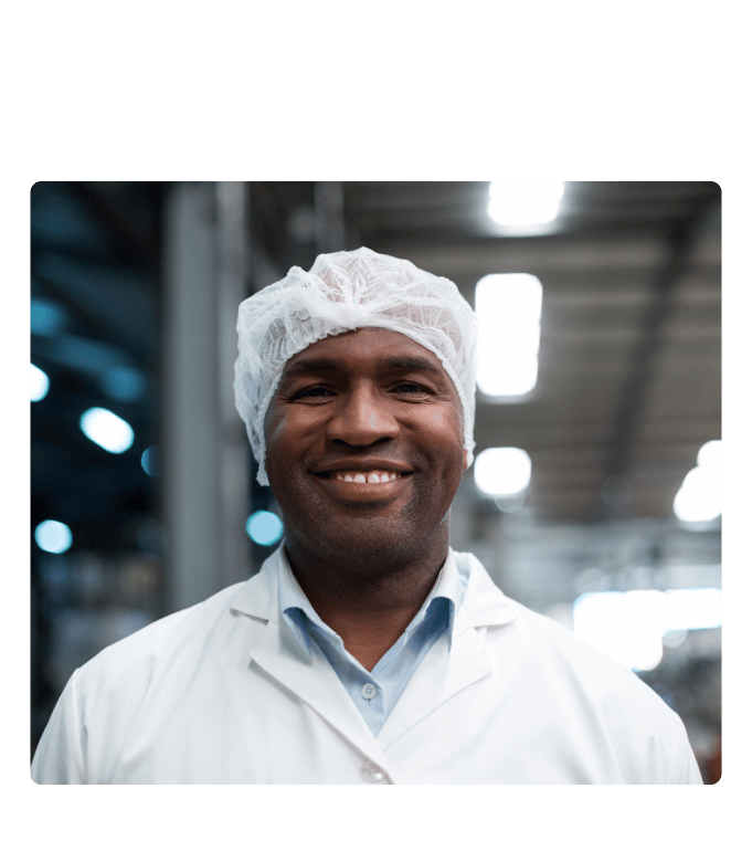 Smiling manufacturing employee in a hairnet and lab coat inside a warehouse facility.