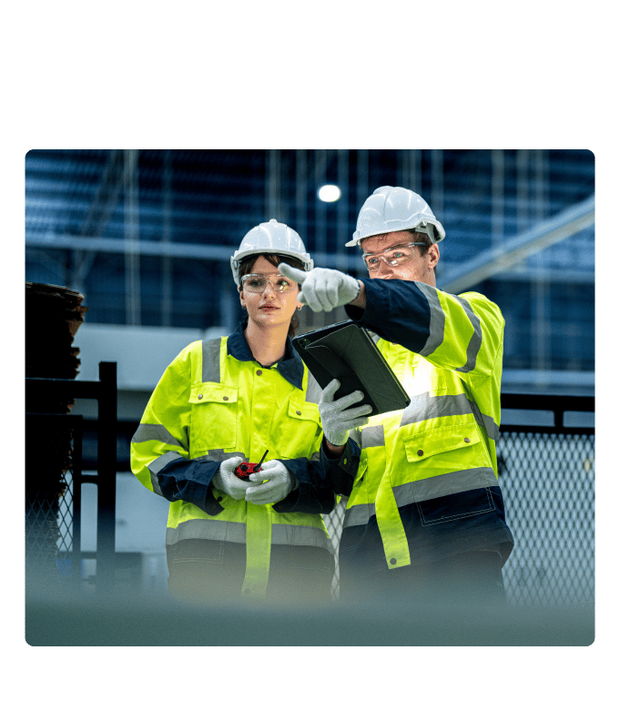 Two industrial workers in safety gear reviewing a tablet on a facility floor.