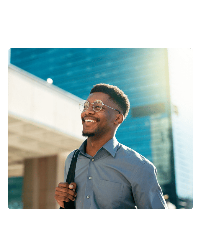 Person wearing glasses and a bag strap smiling outdoors near a glass office building on a sunny day