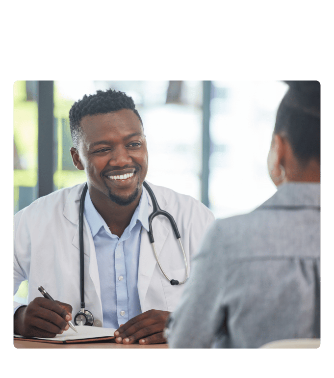 Healthcare provider in a white coat and stethoscope smiling while taking notes with a person in a bright office.