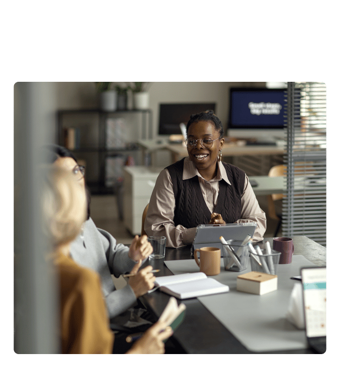 Group of employees in conversation at an office table, one holding a tablet, with notebooks and coffee cups nearby.
