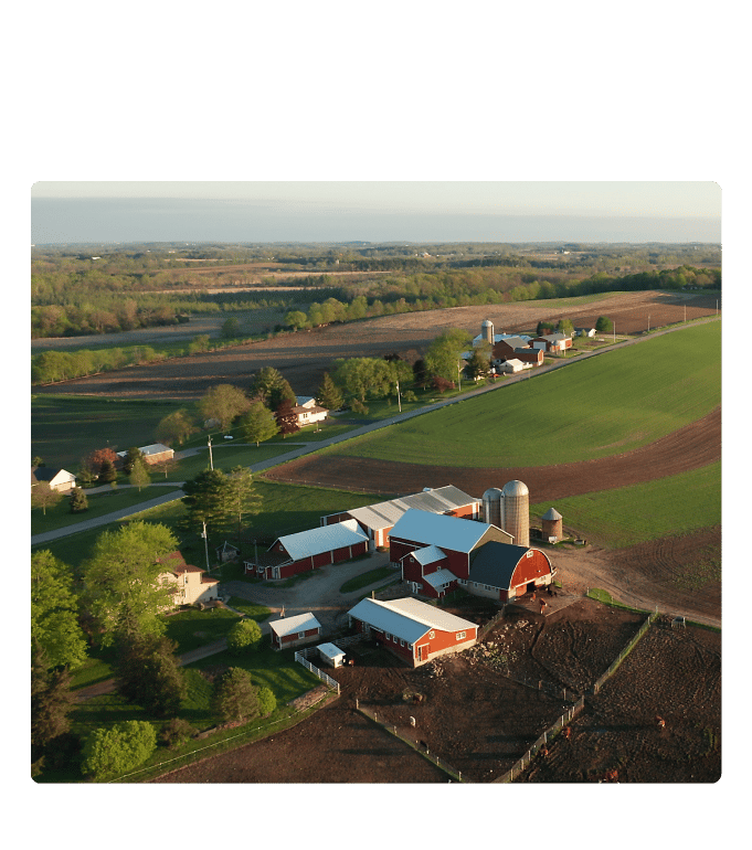 Aerial view of a rural farming community with red barns, a grain silo, and open fields stretching across the countryside.