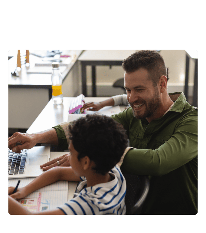 Teacher smiling and pointing at a laptop while working alongside a child at a desk in a classroom setting.