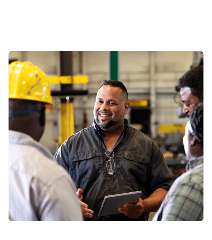 Person holding a tablet smiling while talking with coworkers in an industrial workplace setting.