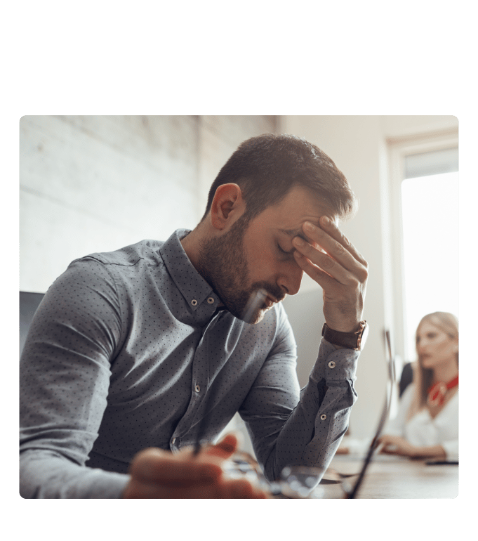 Stressed employee at a desk with head in hand in an office setting, with colleagues visible in the background.