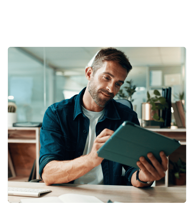 Person reviewing a tablet while seated at an office desk with plants and shelving in the background.