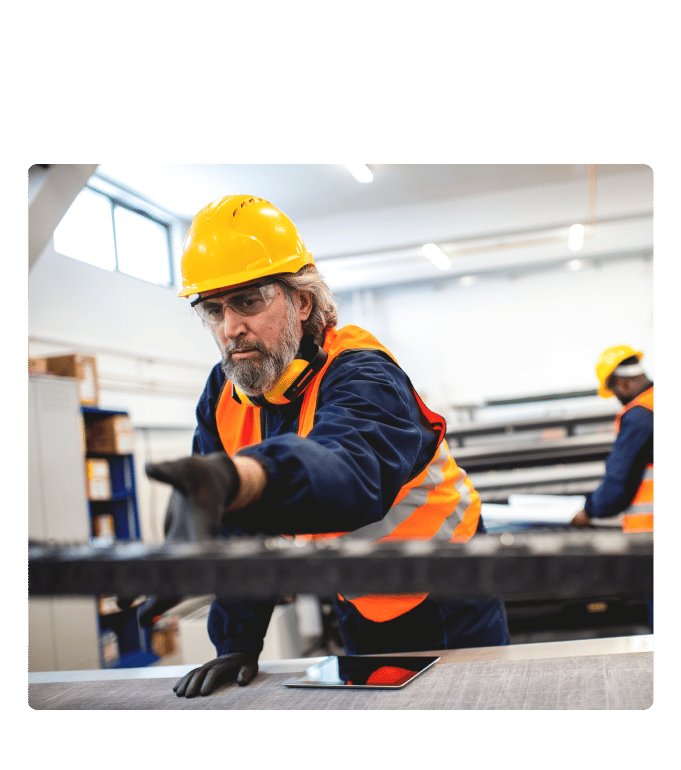 Worker in a hard hat and high-visibility vest handling materials on a factory floor with a tablet nearby