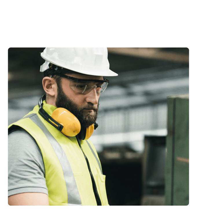 Worker in a hard hat, safety glasses, and high-visibility vest with ear protection in an industrial setting