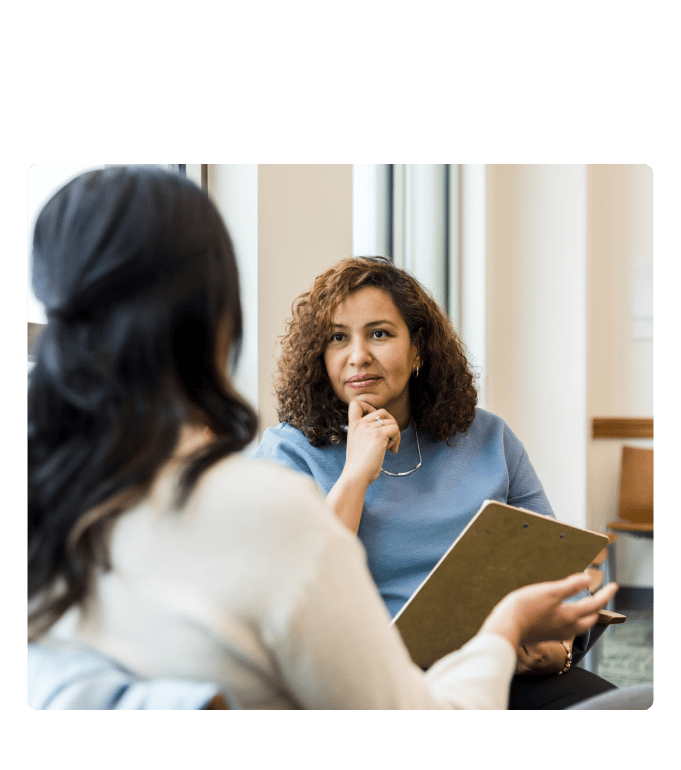 Two people in conversation in an office setting, one holding a clipboard and listening attentively
