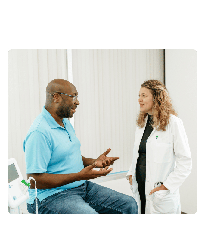 Marathon Health doctor in a white coat speaking with a seated individual in a clinical exam room with medical equipment nearby.