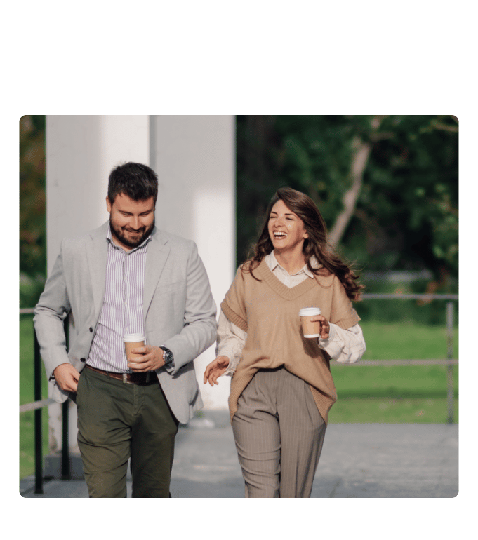 Two employees in good physical health walking outdoors together, smiling and holding takeaway coffee cups, in a park-like setting.