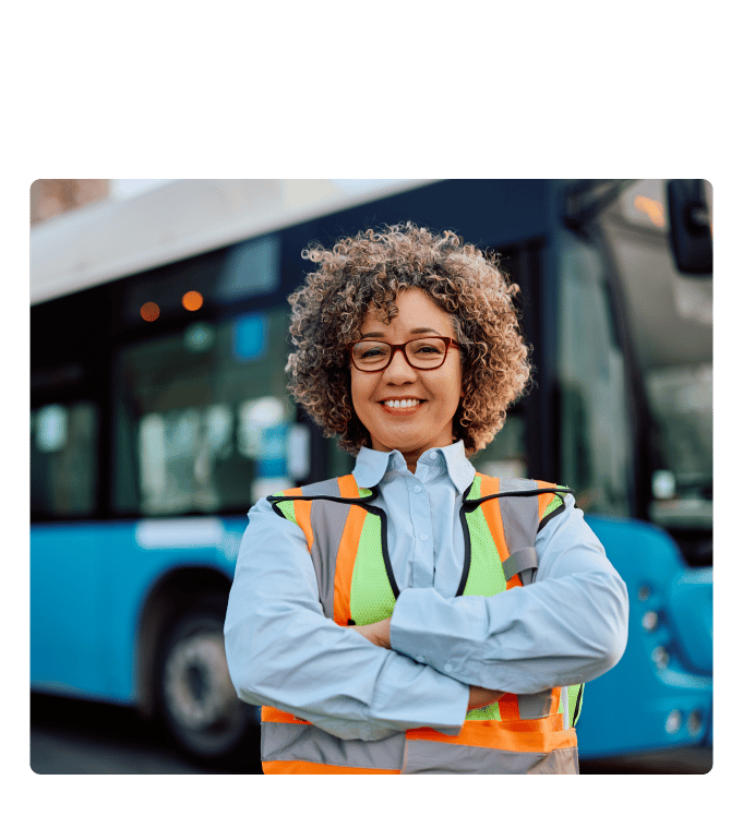 Worker in a high-visibility safety vest standing with arms crossed in front of a transit bus, smiling at the camera