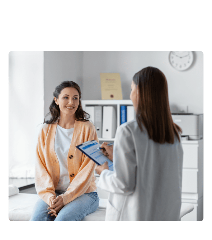 Doctor in a white coat with a clipboard speaking with a patient seated on an exam table.