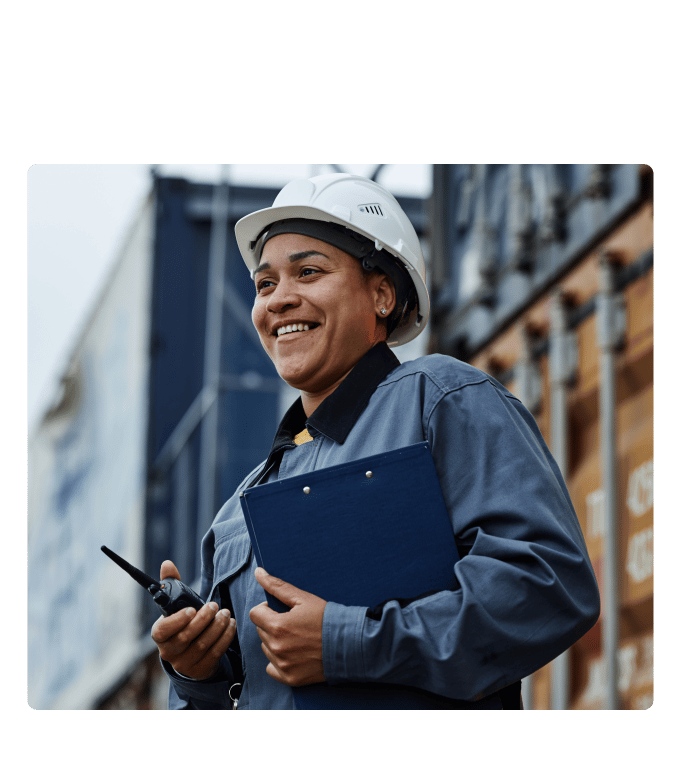 Union worker in a hard hat and work jacket holding a clipboard and radio at an industrial site.