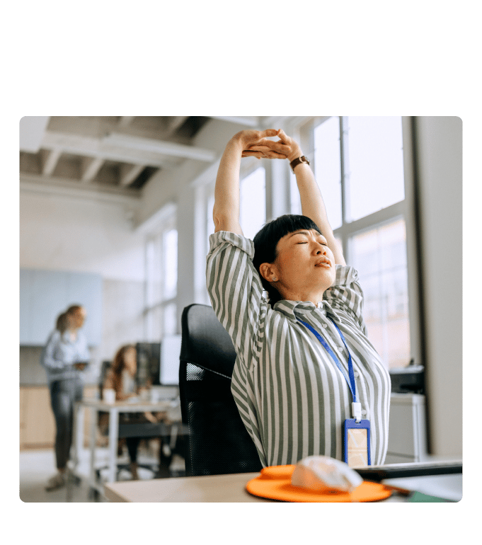Employee stretching arms overhead at a workplace desk, with coworkers and bright windows visible in the background.