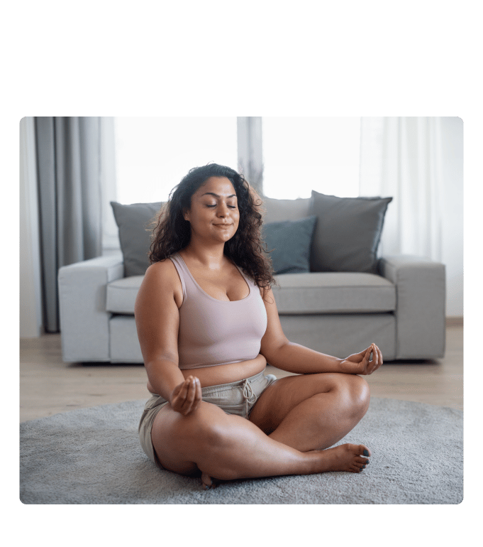 Person sitting cross-legged on a living room floor in a meditation pose with eyes closed