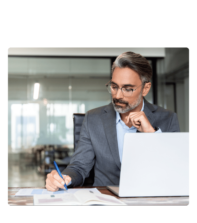 Person in a suit taking notes on paper while reviewing a laptop at a desk in an office setting