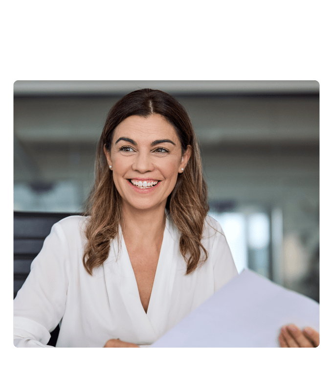 Teacher in a white blouse smiling and holding documents while seated at an office desk.