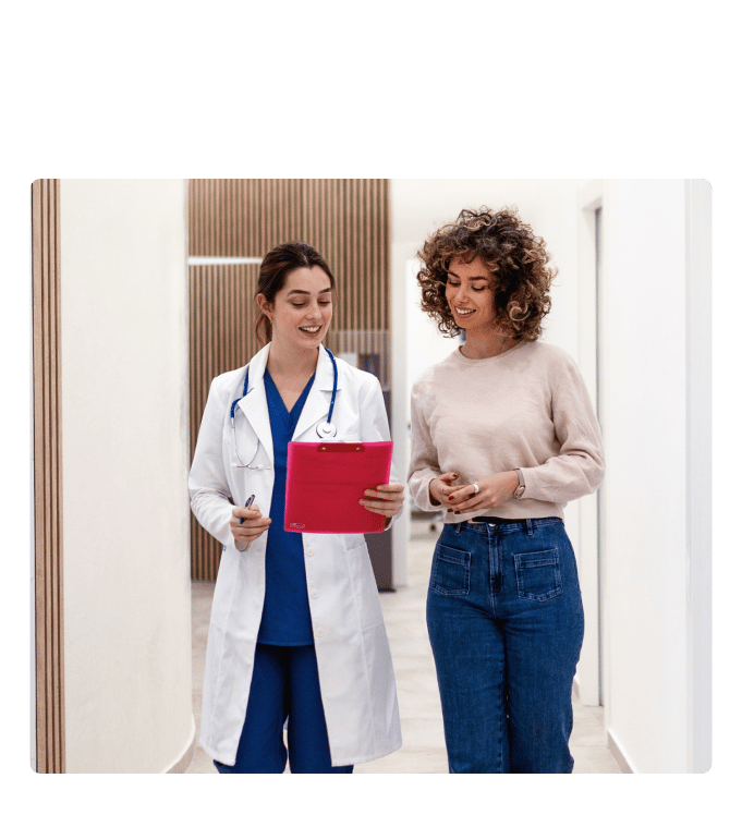 Healthcare provider in a white coat and stethoscope walking and reviewing a clipboard with a person in a clinic hallway