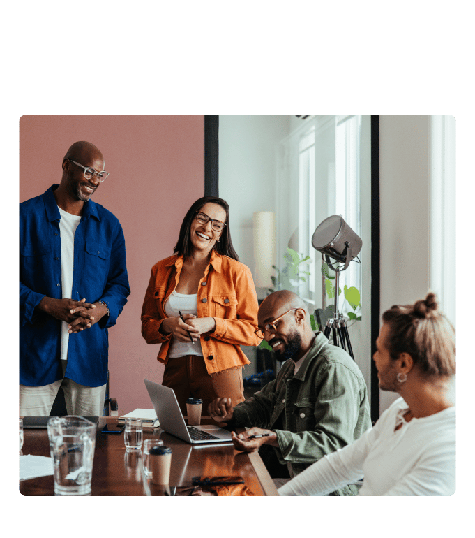Four employees laughing and conversing around a conference table in a modern office setting.