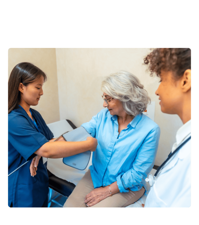 Medical assistant in scrubs applying a blood pressure cuff on a person's arm, observed by a clinician with a stethoscope