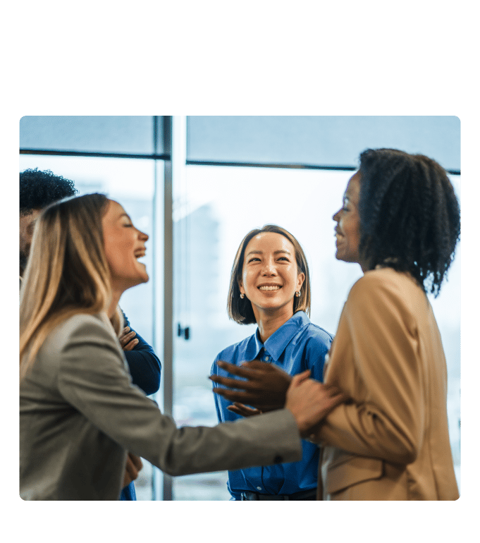 Three employees smiling and conversing in a bright office with large windows.
