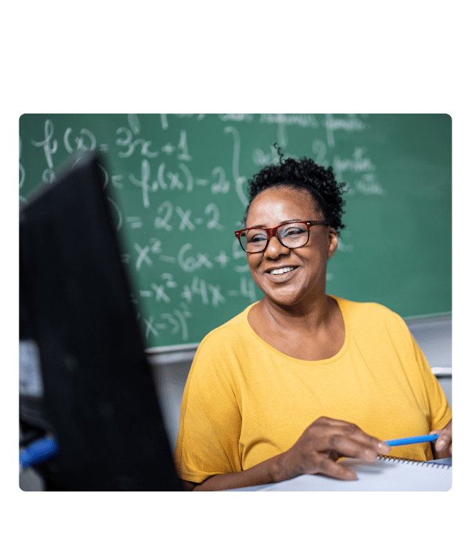 Teacher smiling at a computer screen while writing notes, seated in front of a chalkboard covered in math equations.