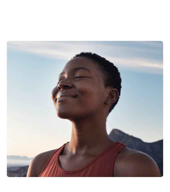 Person practicing mindfulness outdoors, eyes closed, relaxed expression, rocky landscape and blue sky behind them.