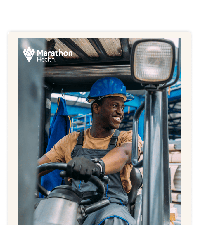 A smiling worker in a blue hard hat and overalls operates heavy equipment inside an industrial facility