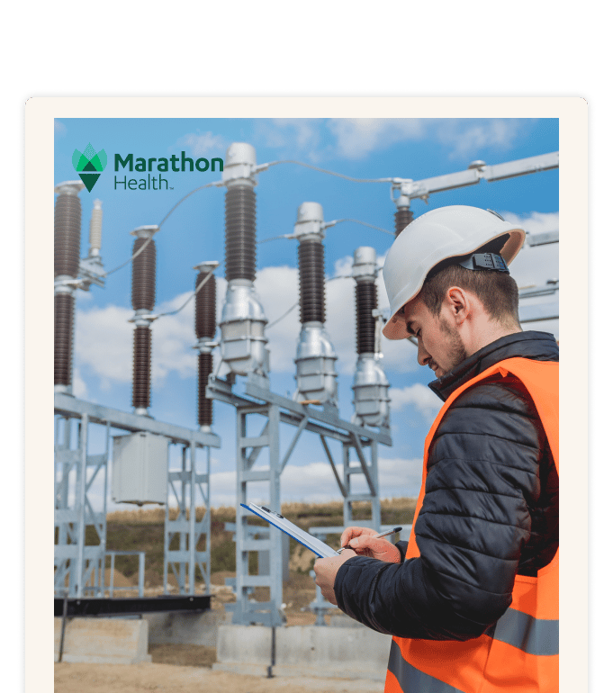 A worker in an orange safety vest and white hard hat writes on a clipboard at an industrial electrical substation