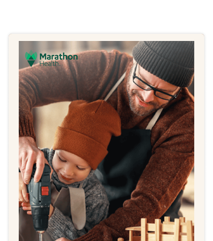 A bearded person wearing glasses and a knit hat guides a child using a power drill at a wooden workbench
