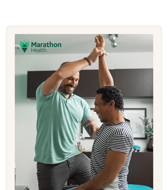 Two smiling people indoors, one guiding the other's arm overhead during a hands-on session, Marathon Health logo visible