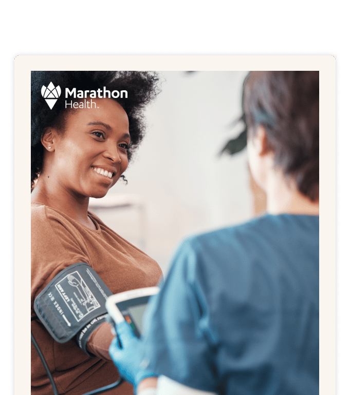 A smiling person has a blood pressure cuff on their arm while a healthcare provider in scrubs assists during a clinic visit