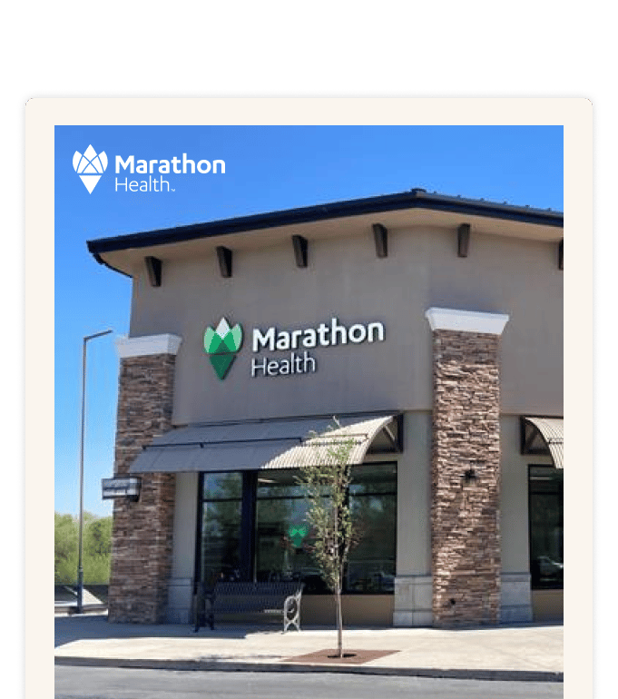 Exterior of a Marathon Health network health center with stone columns, large windows, and a small tree out front under a clear blue sky.