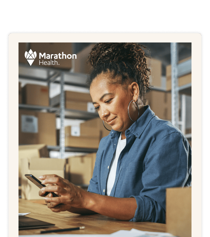 A person in a denim shirt uses a smartphone at a desk surrounded by cardboard boxes on warehouse shelves