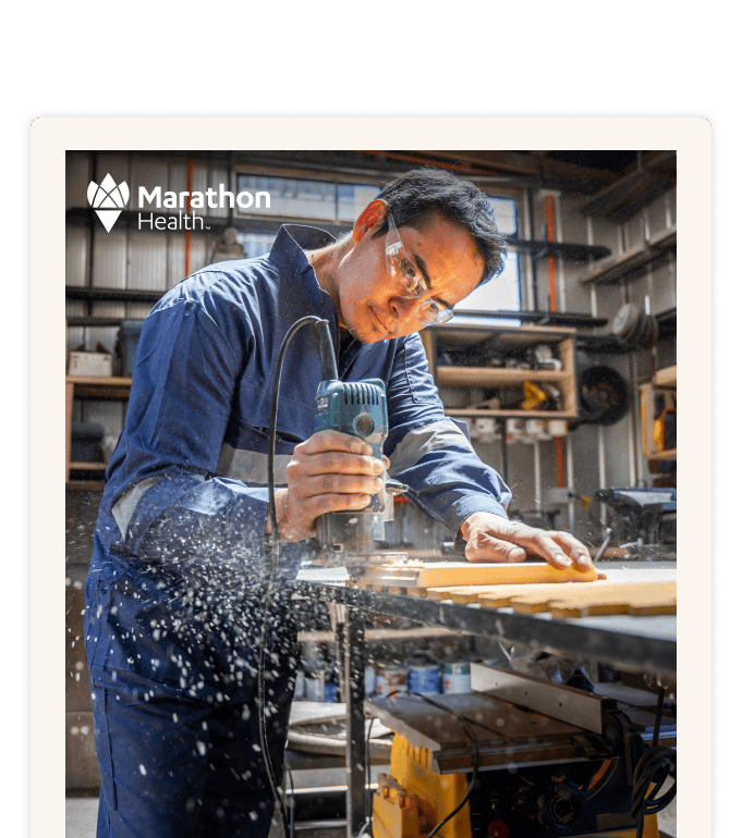 A person in blue coveralls and safety glasses uses a power tool on wood in a workshop with sawdust flying