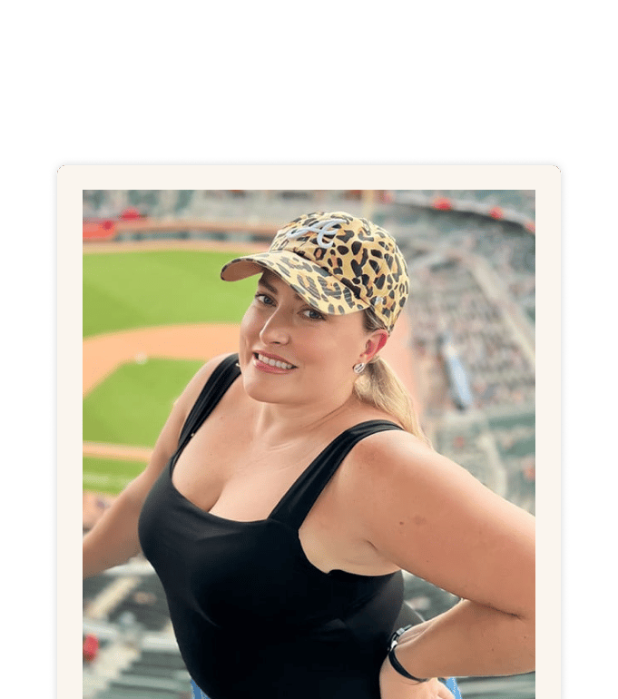 Rachel Strouse smiling at a baseball stadium, wearing a black tank top and leopard-print cap