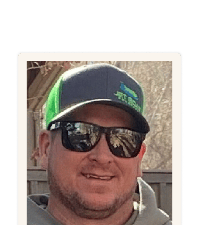 Cody smiling outdoors in a green and gray snapback cap and mirrored sunglasses near a wooden fence
