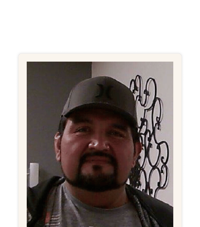 Carlos wearing a dark baseball cap and casual jacket, standing indoors near a decorative metal wall piece