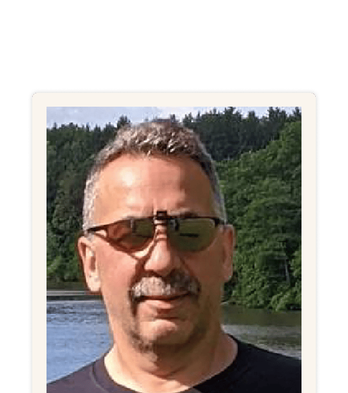 Andrew wearing sunglasses and a dark t-shirt, standing outdoors near a lake with forested hills behind him