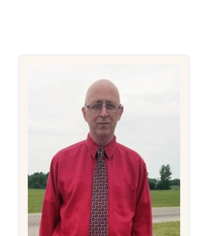 Brian Wiley standing outdoors in a red dress shirt and patterned tie, with green fields and cloudy sky behind him