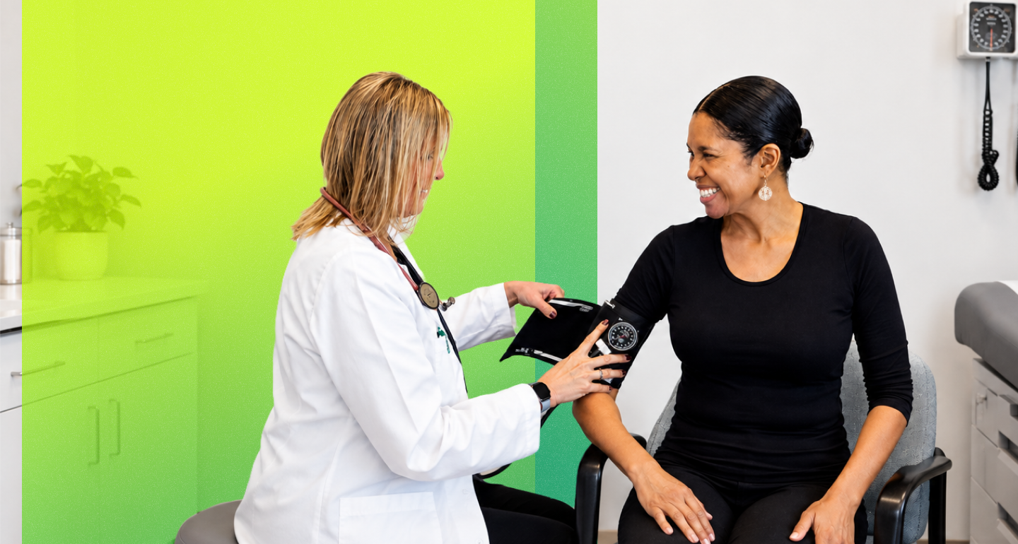 A healthcare provider in a white coat uses a blood pressure cuff on a smiling person seated in a clinical room