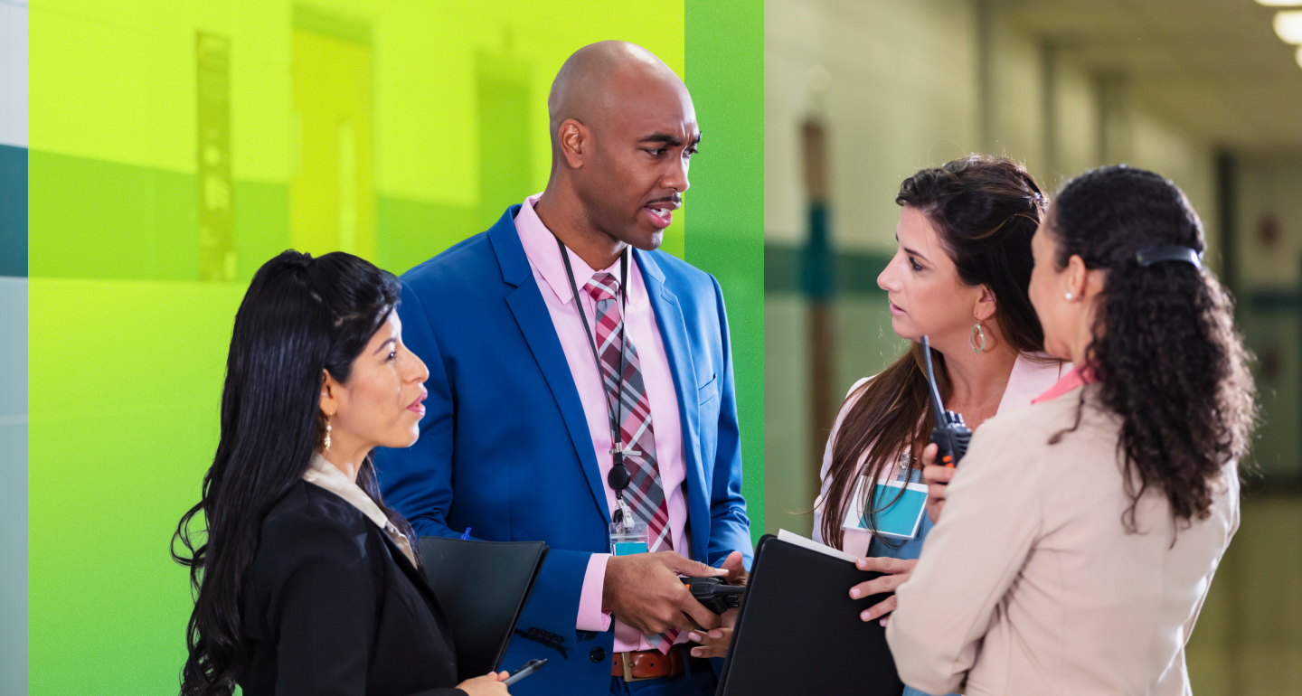 Four professionals in business attire having a discussion in a hallway, holding clipboards and a walkie-talkie