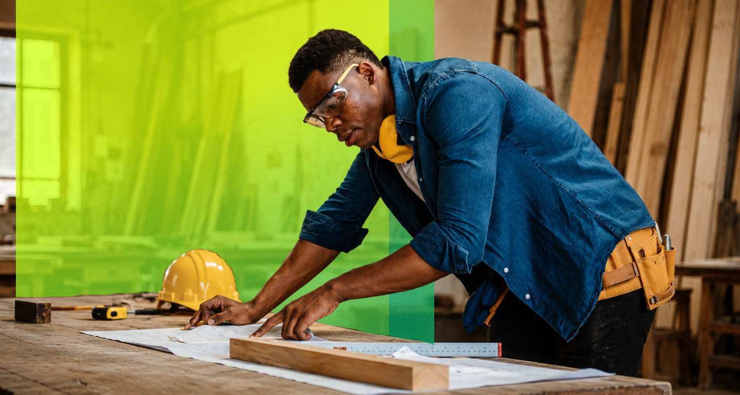 A person in a denim shirt, tool belt, safety glasses, and ear protection reviews blueprints on a workbench beside a hard hat