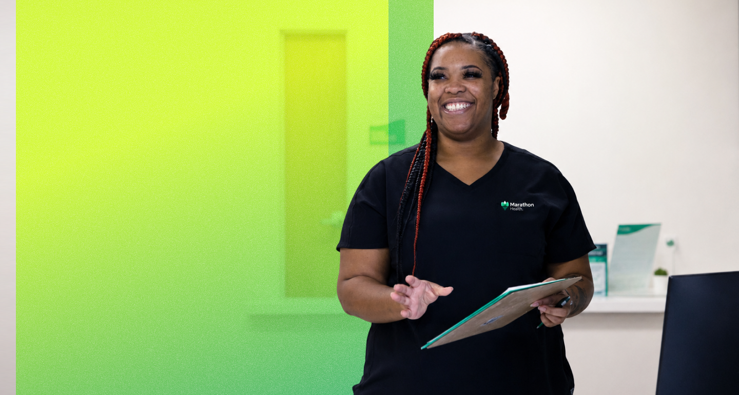 A smiling Marathon Health provider in black scrubs holds a clipboard inside a bright clinic with a green accent wall